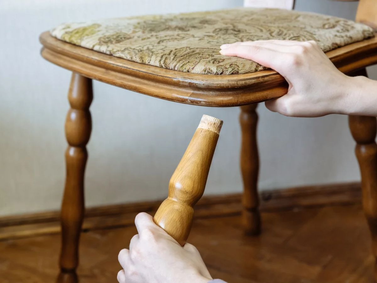A person repairs a wooden stool, holding a leg with a worn edge while stabilizing the seat with their other hand.