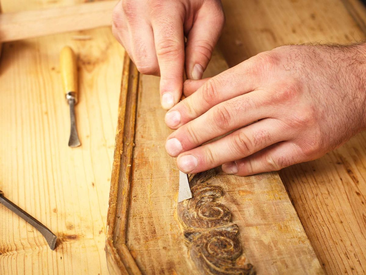 A craftsman uses a chisel to carve detailed designs into a wooden panel, showcasing his woodworking skills at a workbench.