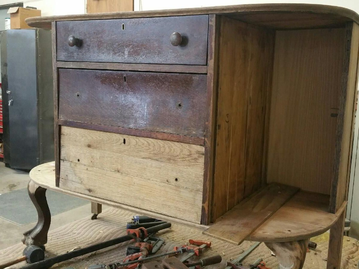A wooden cabinet with two drawers is under restoration in a workshop, showcasing exposed wood and surrounded by various tools.