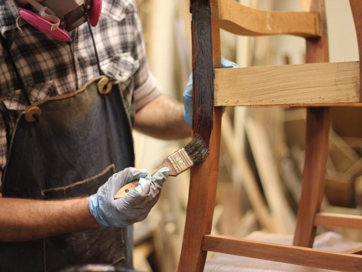 A craftsman applies wood stain to a chair leg, wearing gloves and a mask, surrounded by various woodworking tools.