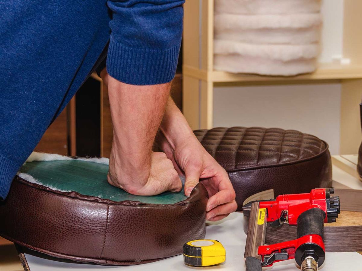 A person is reupholstering a circular brown cushion, using a staple gun and measuring tape on a work surface.