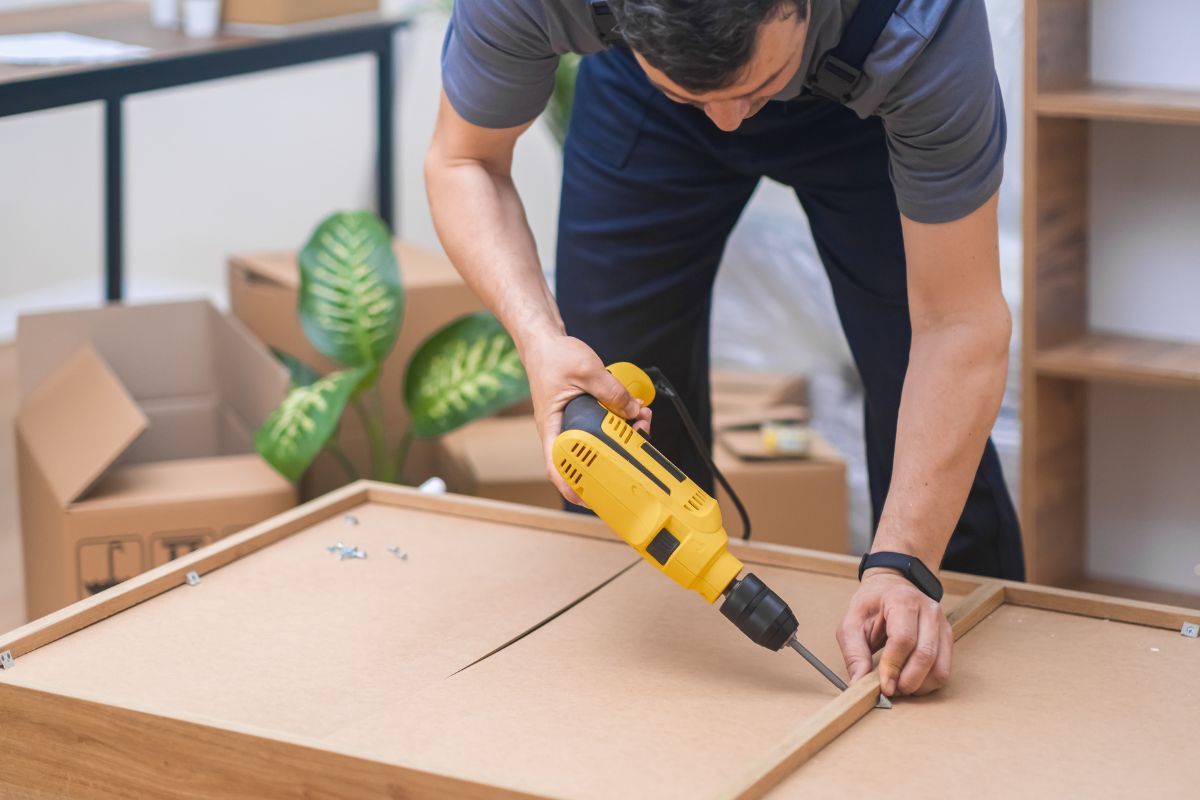 A person using a yellow power drill to assemble furniture, surrounded by cardboard boxes and indoor plants.