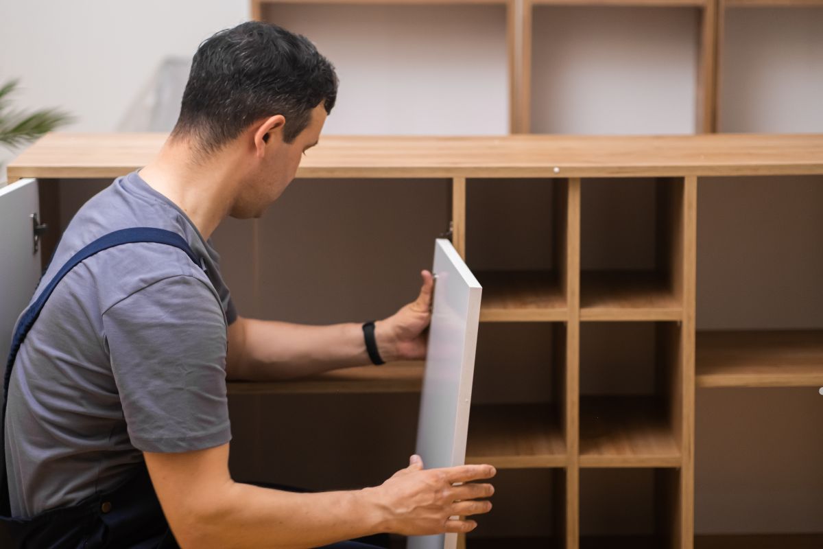A person in a gray T-shirt is installing a white cupboard door on a wooden storage unit with square compartments.