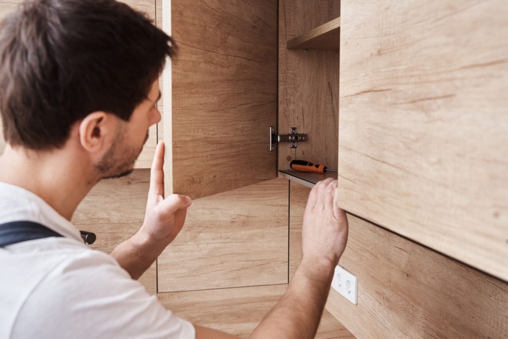 A person examines the inside of a wooden cabinet, revealing a screwdriver on the shelf. The cabinetry features a natural wood finish.