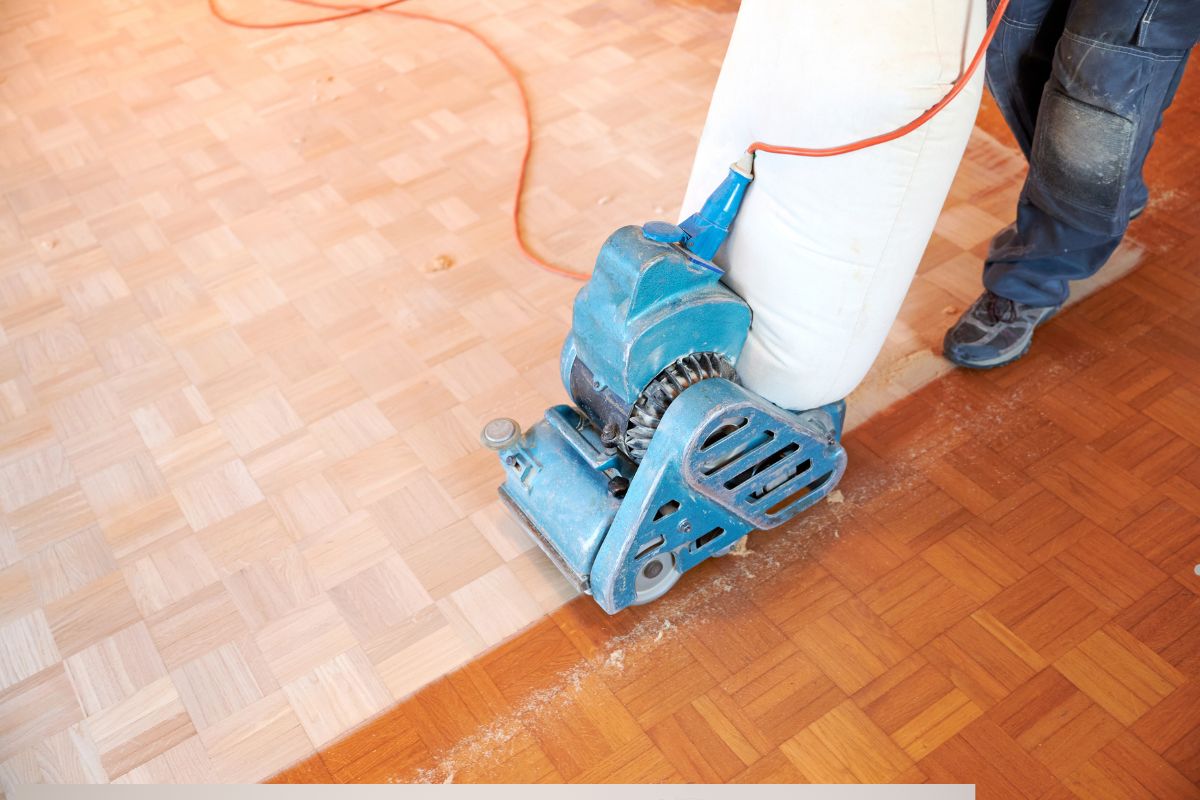 A person using a blue floor sanding machine on wooden parquet flooring, with visible sawdust and a power cord in the background.
